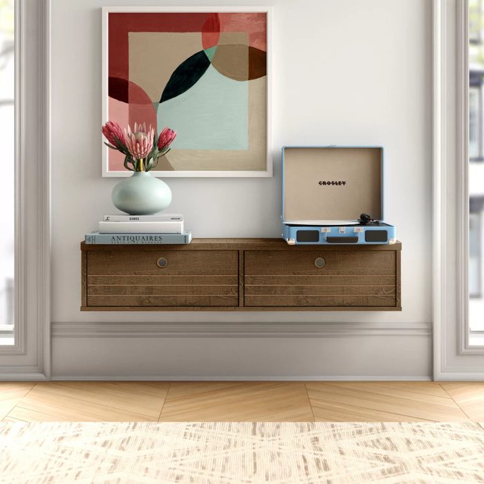 A wall-mounted wooden shelf holds a decorative vase with flowers and a vintage record player, accompanied by stacked books, against a light-colored wall.