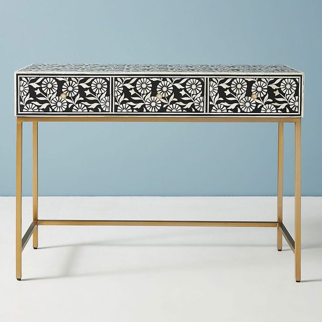 A decorative console table with floral patterns on three drawers, supported by a sleek gold frame, stands against a light blue wall.