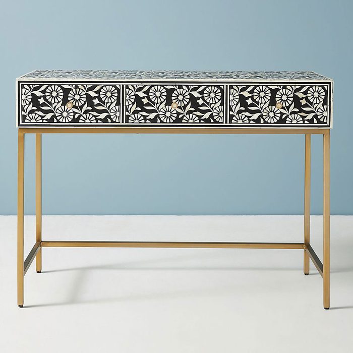 A decorative console table with floral patterns on three drawers, supported by a sleek gold frame, stands against a light blue wall.
