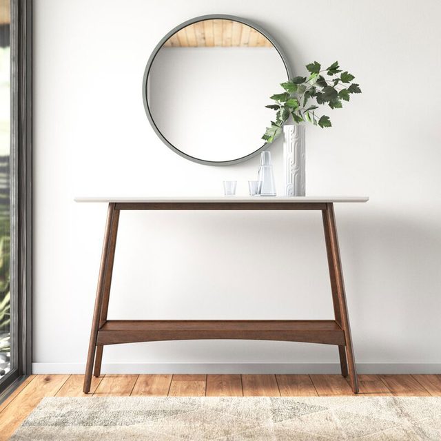 A wooden table with a white top displays a vase of greenery and glassware, situated in a bright room near a mirror and a window.