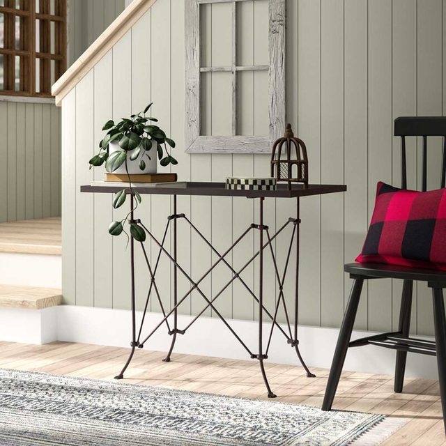 A metal table supports a plant and decorative items in a cozy, light-colored room with wooden stairs, a patterned rug, and a black chair.