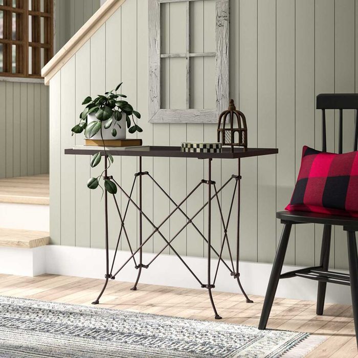 A metal table supports a plant and decorative items in a cozy, light-colored room with wooden stairs, a patterned rug, and a black chair.