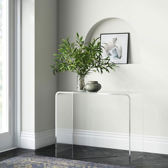 A clear, modern table holds a potted plant and decorative items in a bright corner with large windows and a muted wall, featuring framed art above.