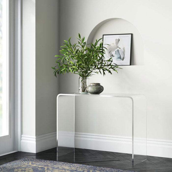 A clear, modern table holds a potted plant and decorative items in a bright corner with large windows and a muted wall, featuring framed art above.