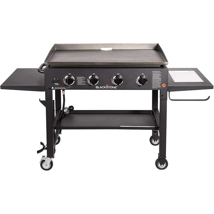 A large, flat-top griddle with four knobs on the front, placed on a wheeled metal cart, designed for outdoor cooking.
