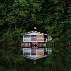 A small, perched house reflects off calm water, surrounded by dense greenery, creating a serene, picturesque setting.