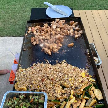 Grilled chicken bites sizzle on a hot cooking surface alongside fried rice and vegetables, set outdoors on a wooden deck with grass nearby.