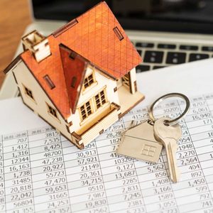 A miniature house sits on financial documents with keys attached to a keyring nearby, suggesting a focus on real estate transactions or investments.