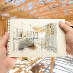 A hand holds an illustrated bathroom design on a notebook. The background shows a construction site with exposed wooden beams and unfinished structures.