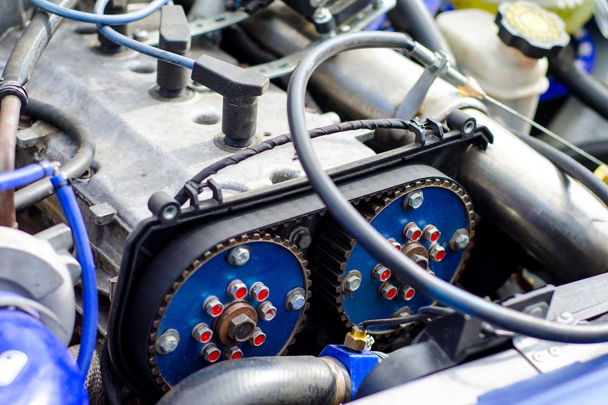 An engine component features blue gears connected by a black belt, surrounded by metallic parts and hoses in an automotive setting.