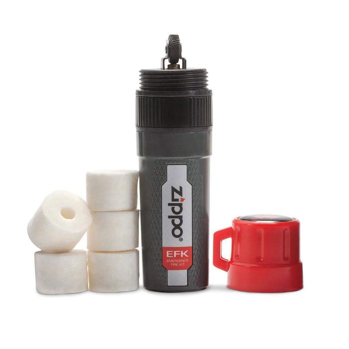 A cylindrical container holds fire-starting material, surrounded by white wax cubes. A red cap rests beside the container, indicating a fire kit for emergencies.