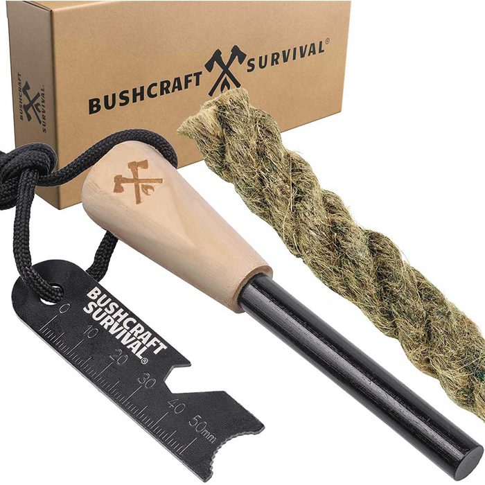 A fire-starting kit includes a wooden striker, a black metal scraper, and a twisted fiber bundle, all positioned beside a branded cardboard box.