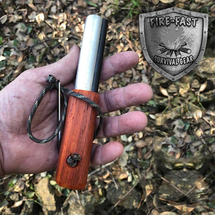 A hand holds a metal and wooden fire starter tool amidst a forest floor covered in leaves, suggesting a survival or outdoor setting.