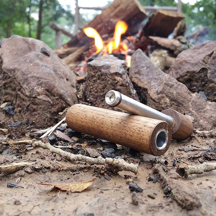 Two cylindrical objects, one wooden and one metallic, rest on the dirt near a fire, surrounded by rocks and scattered twigs in a natural setting.