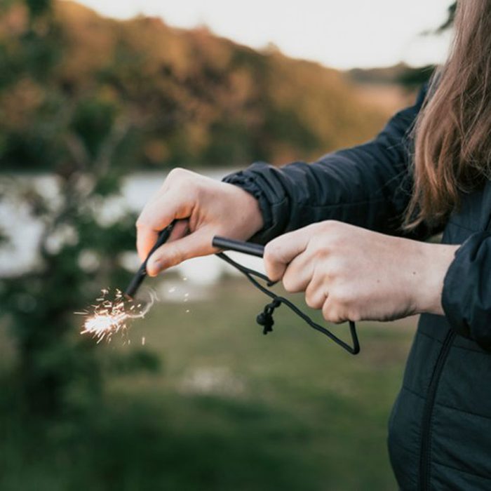 A person strikes a fire starter, producing sparks, in a natural outdoor setting with greenery and water in the background.