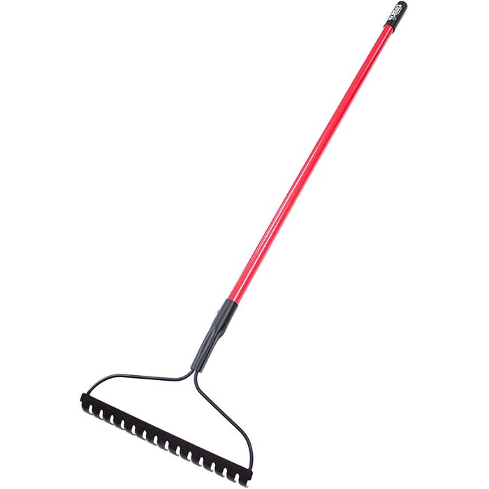 A long red rake with a curved, black metal head and multiple tines rests vertically, designed for leveling soil or collecting debris in outdoor areas.