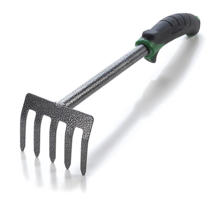 A metal gardening hand rake with a textured grip is positioned on a white surface, used for loosening soil or gathering debris in a garden.