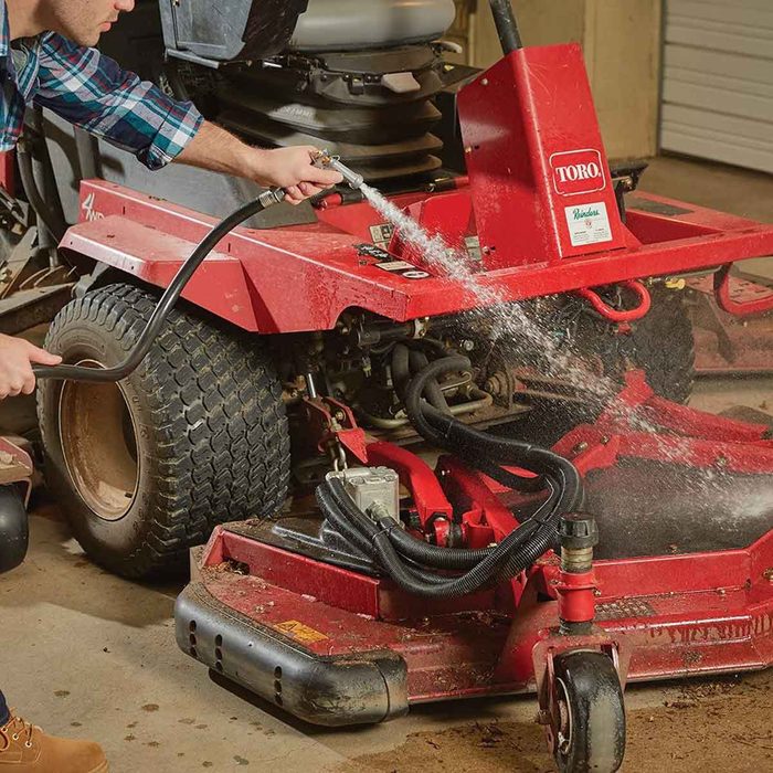 A person washes a red lawn mower using a hose, spraying water on its undercarriage in a garage environment.