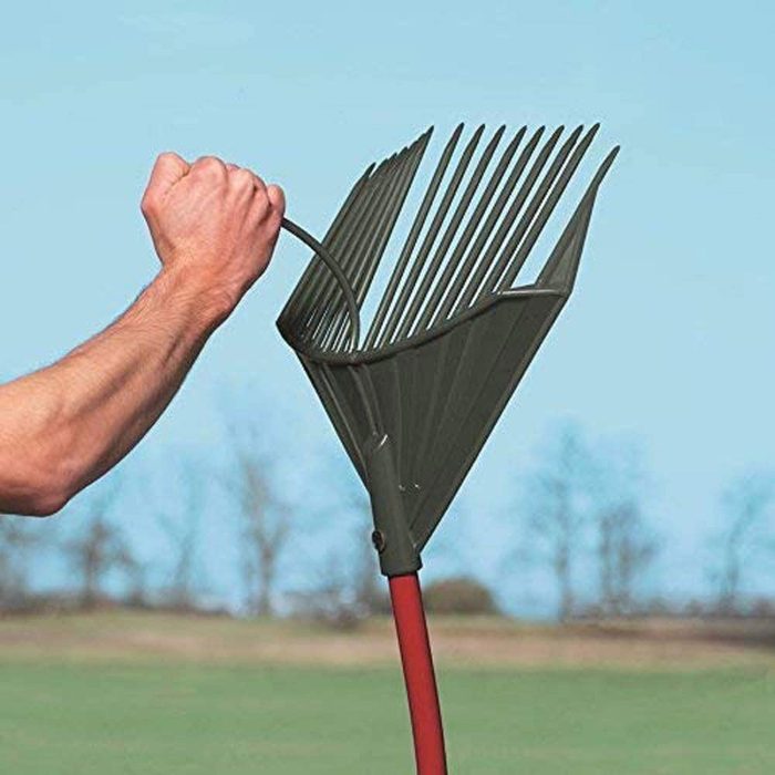 A hand grips a green rake, preparing to gather material. The background features a clear sky and distant trees, suggesting an outdoor setting.