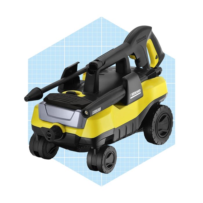 A compact, yellow and black pressure washer sits on four wheels, equipped with a spray lance and nozzle, against a light grid background.