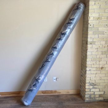 A rolled rug leans against a beige wall corner in a room with wooden flooring, featuring a small electrical outlet nearby.