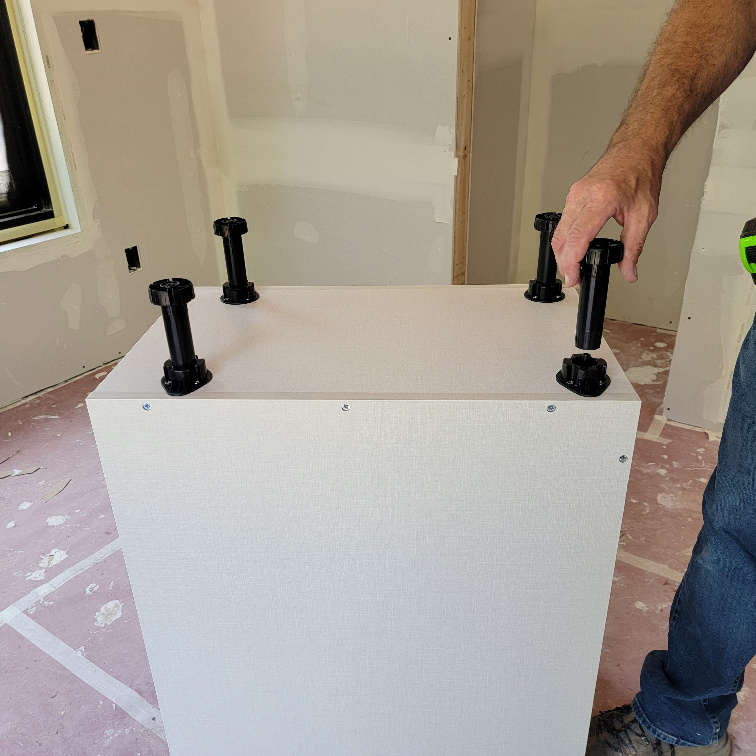 A person adjusts black support legs on a light-colored box in a partially finished room with drywall and exposed framing.