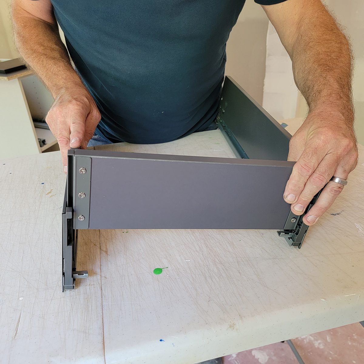 A person assembles a gray drawer front, securing it to a metal frame on a worktable in a partially constructed room.