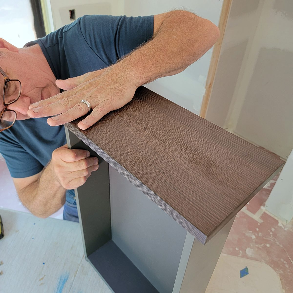 A person is closely inspecting the top of a wooden shelf while adjusting its position in an unfinished interior space.