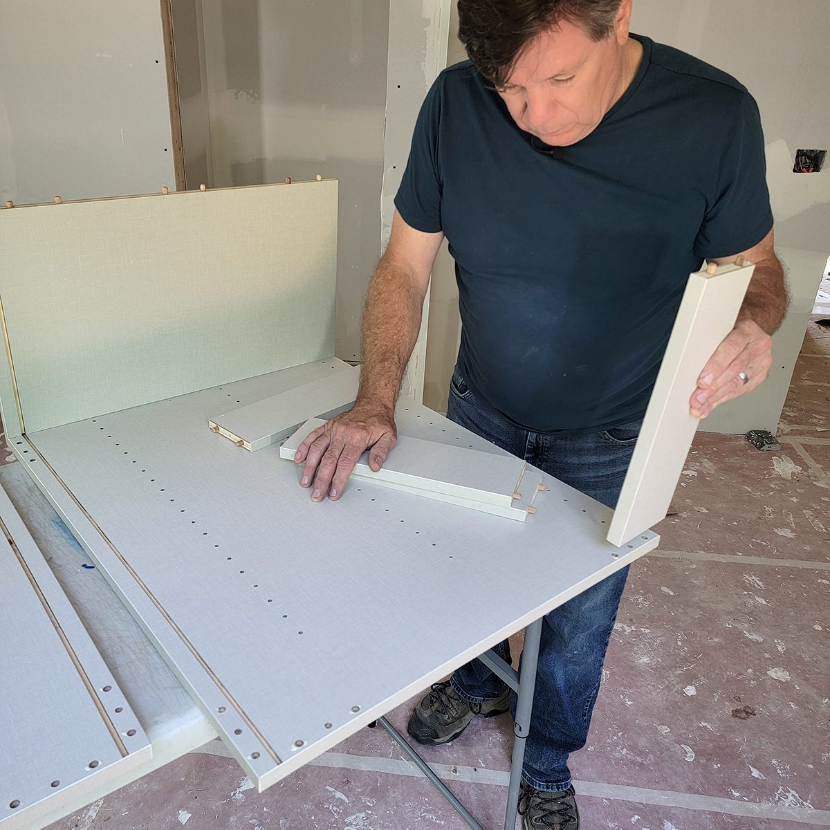 A man assembles furniture parts by aligning them on a table in a partially constructed room with drywall and a bare floor.