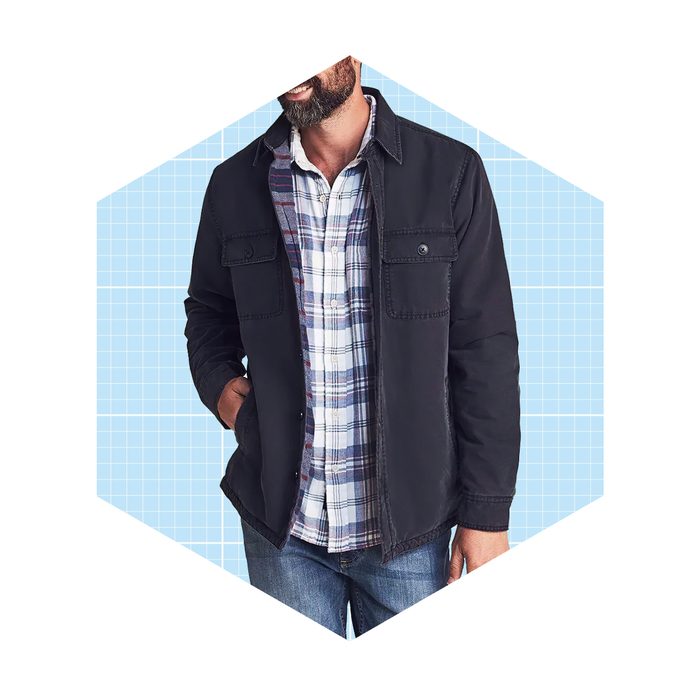 A man wears a black jacket over a plaid shirt, standing casually against a light blue grid background.