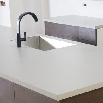 A black faucet is positioned over a square sink on a light-colored countertop, adjacent to a wooden surface, in an unfinished kitchen space.