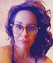 A person with curly hair and glasses gazes at the camera, set against a softly blurred background.