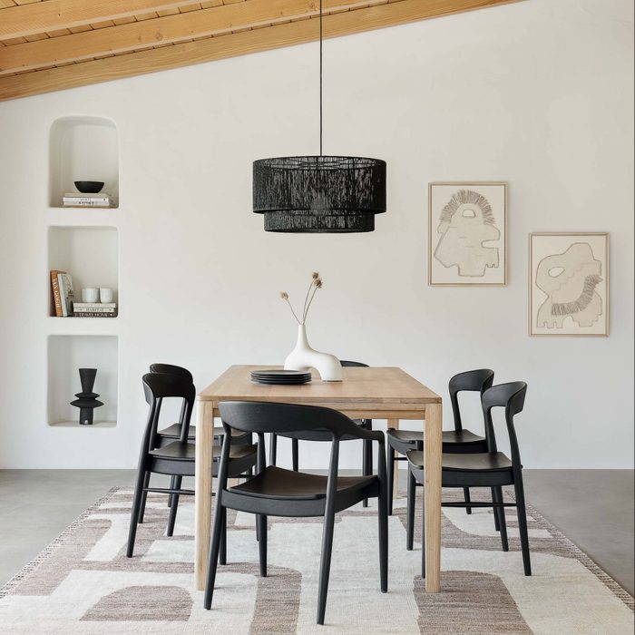 A wooden dining table with six black chairs sits on a patterned rug. Above hangs a pendant light, complementing minimalist artwork on a white wall.
