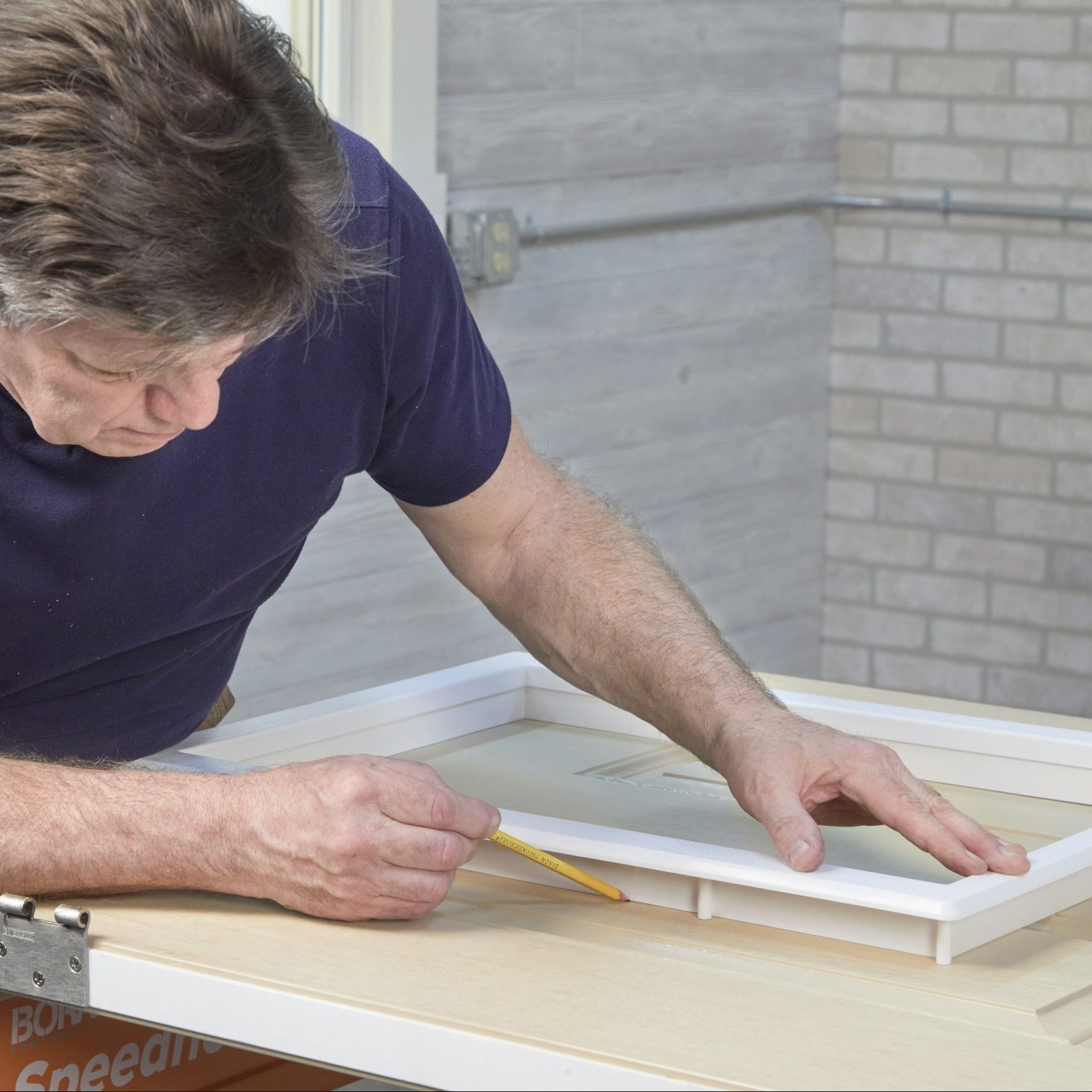 A person measures a frame with a pencil while leaning over a wooden surface in a bright, structured indoor workspace.