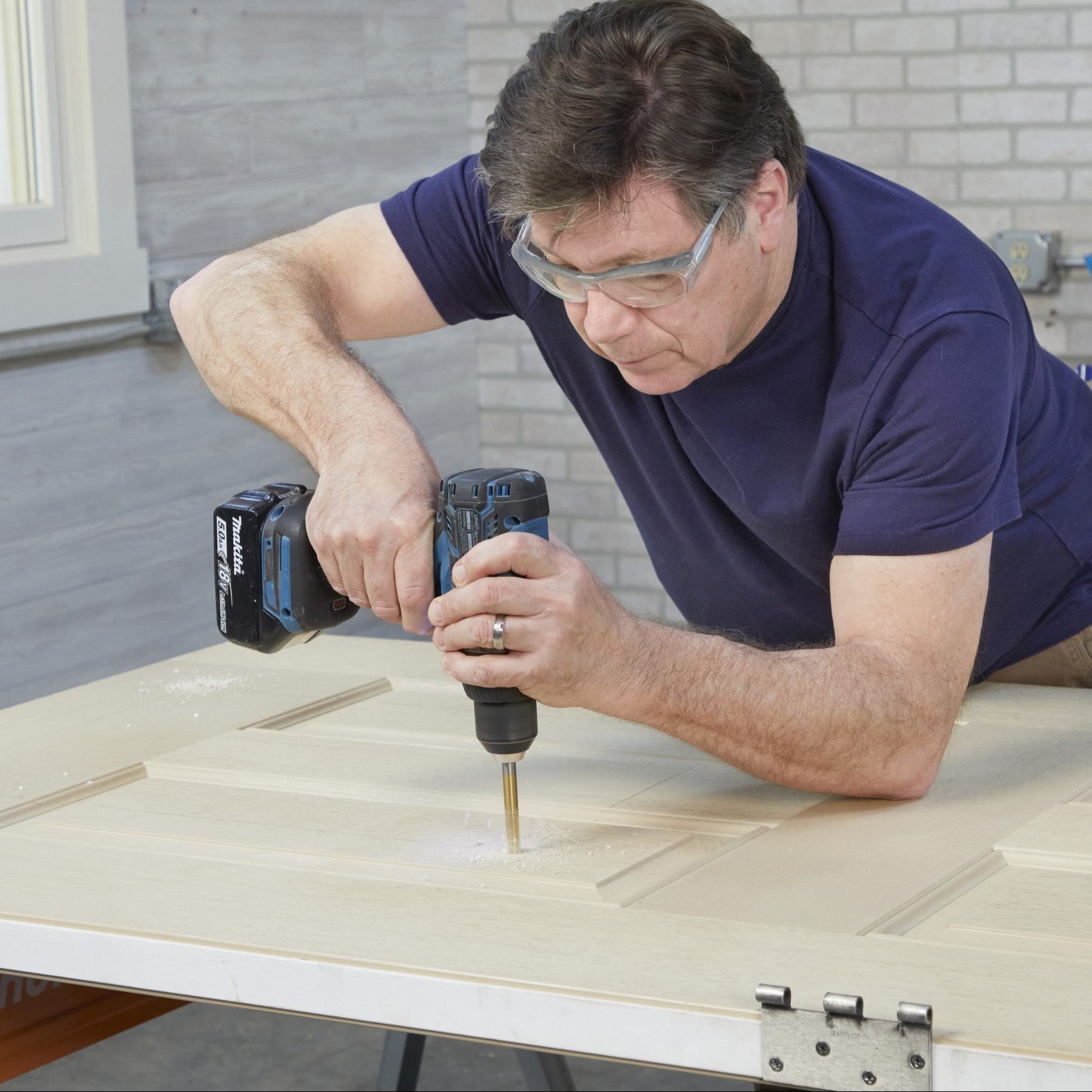 A man drills into a wooden surface, concentrating intently while wearing safety glasses, in a well-lit workshop with brick walls.