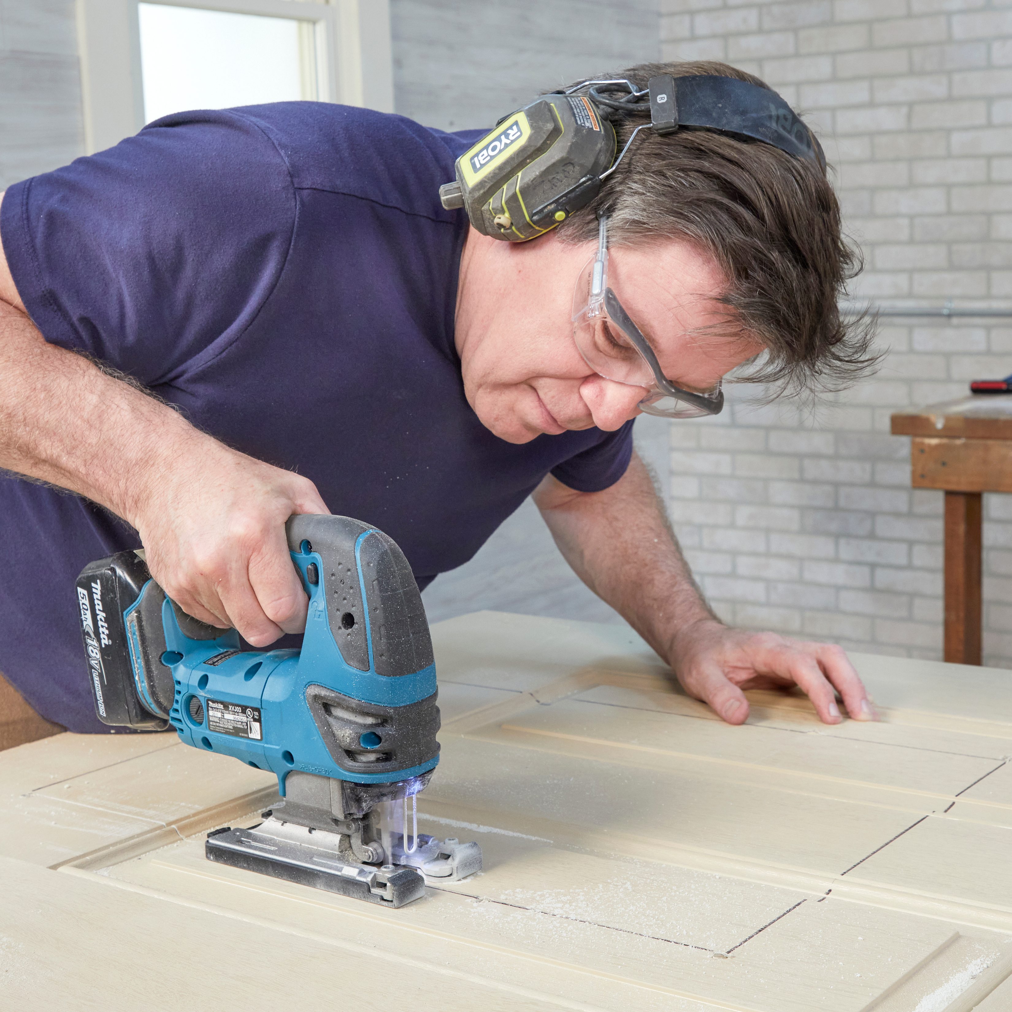 A man operates a jigsaw, cutting into a wooden surface in a well-lit room with a brick wall and a workbench in the background.