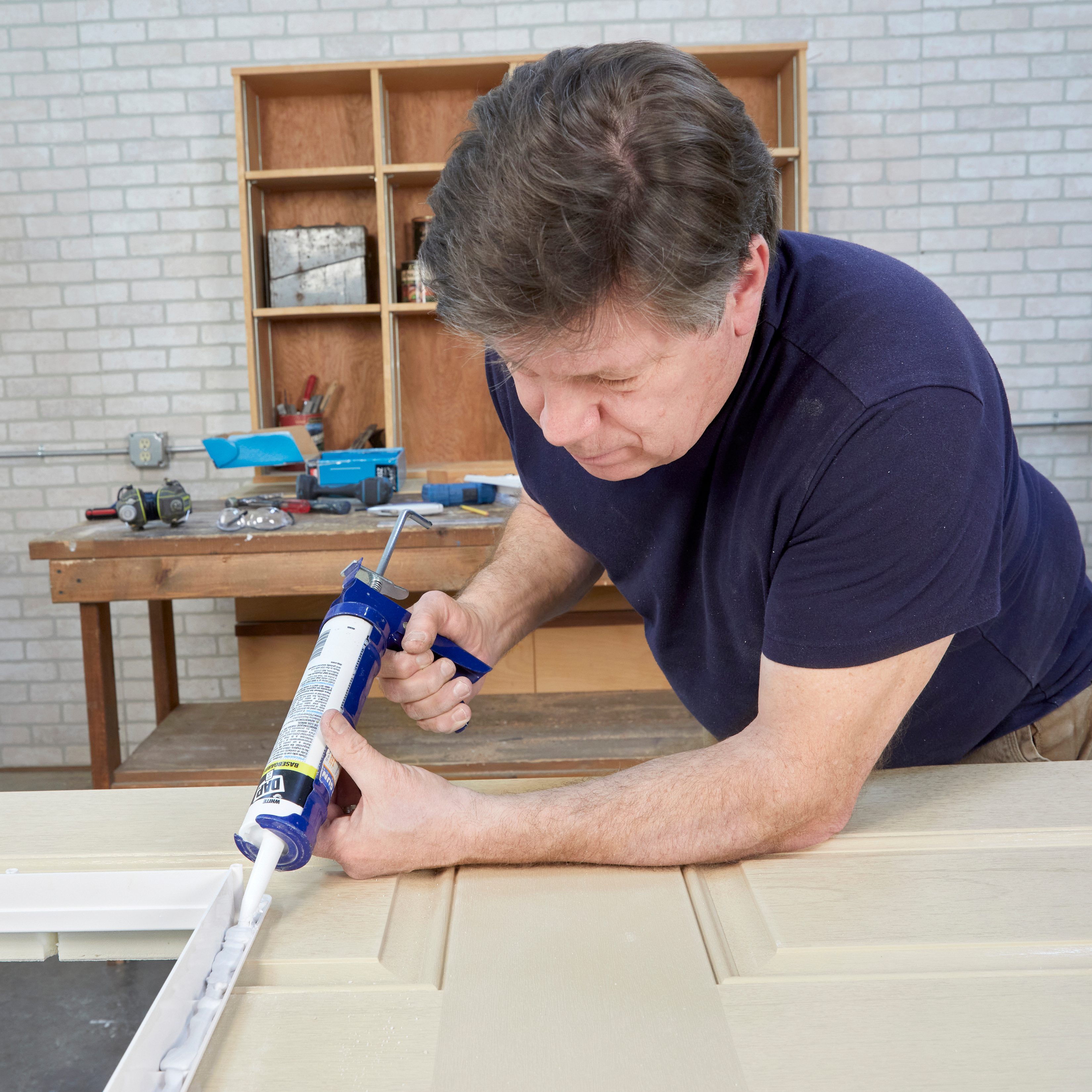 A man applies caulk to a paneled door on a workbench, surrounded by tools and a wooden shelf in a well-lit workshop.