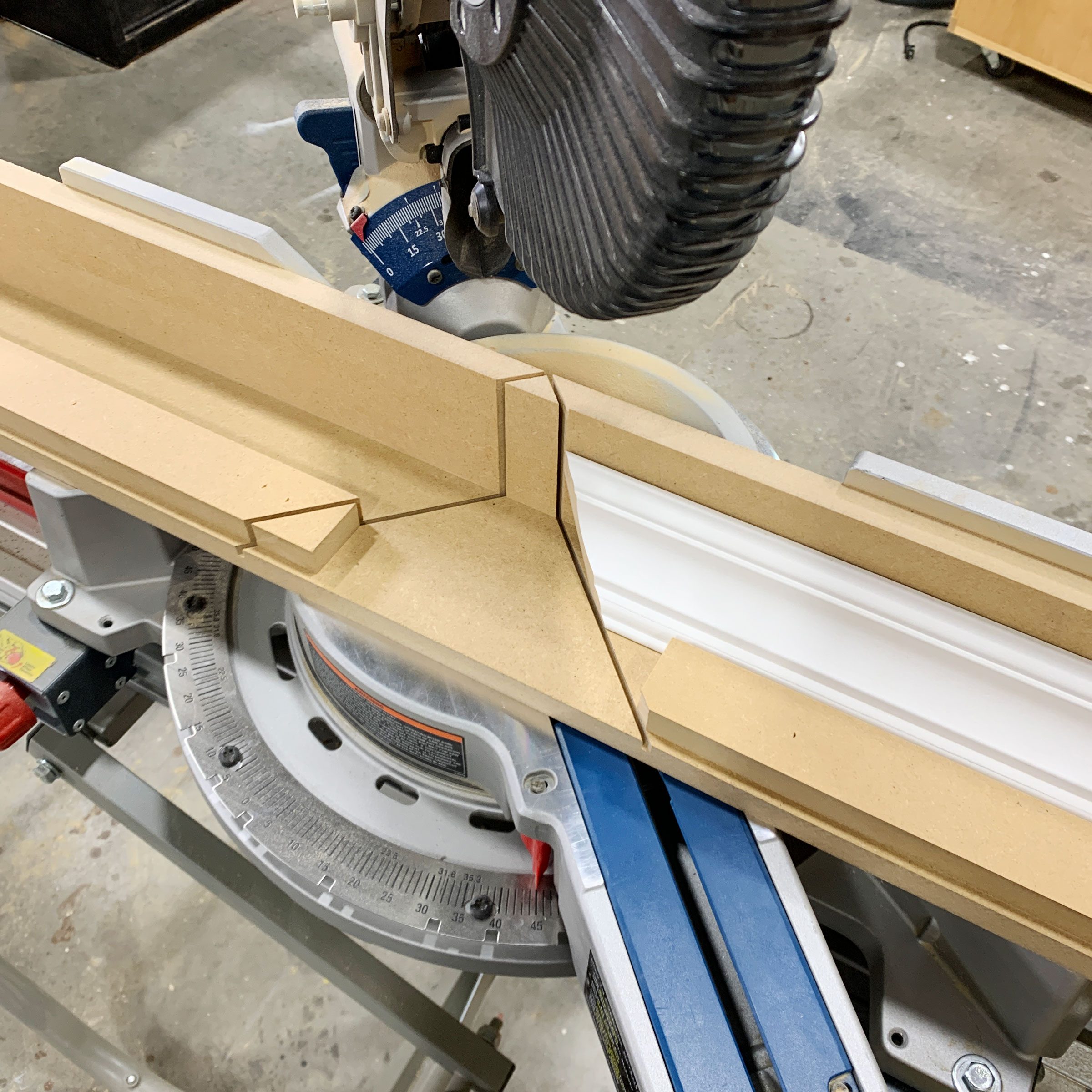 A miter saw with a white trim piece positioned under a saw blade, ready for cutting. The workspace has a concrete floor and wooden surroundings.