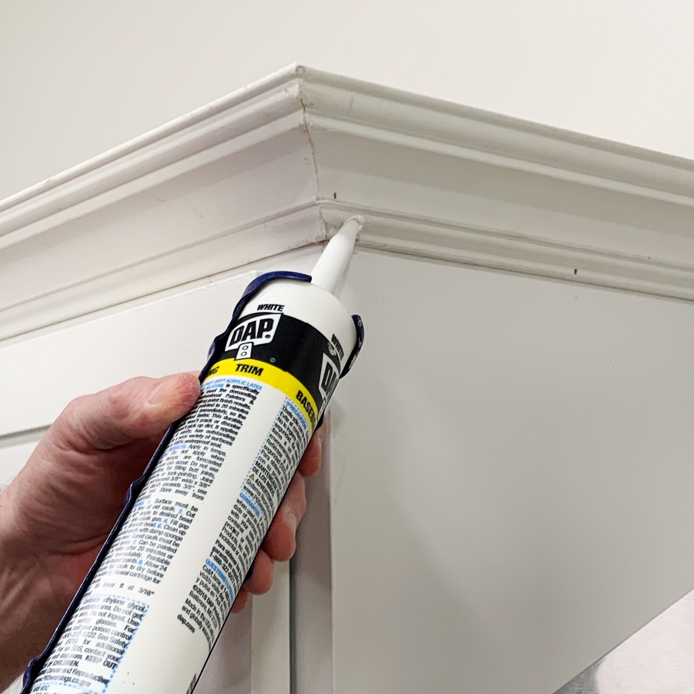 A hand holds a caulking gun, applying sealant to a corner of white trim against a plain wall, focusing on filling gaps for a smooth finish.