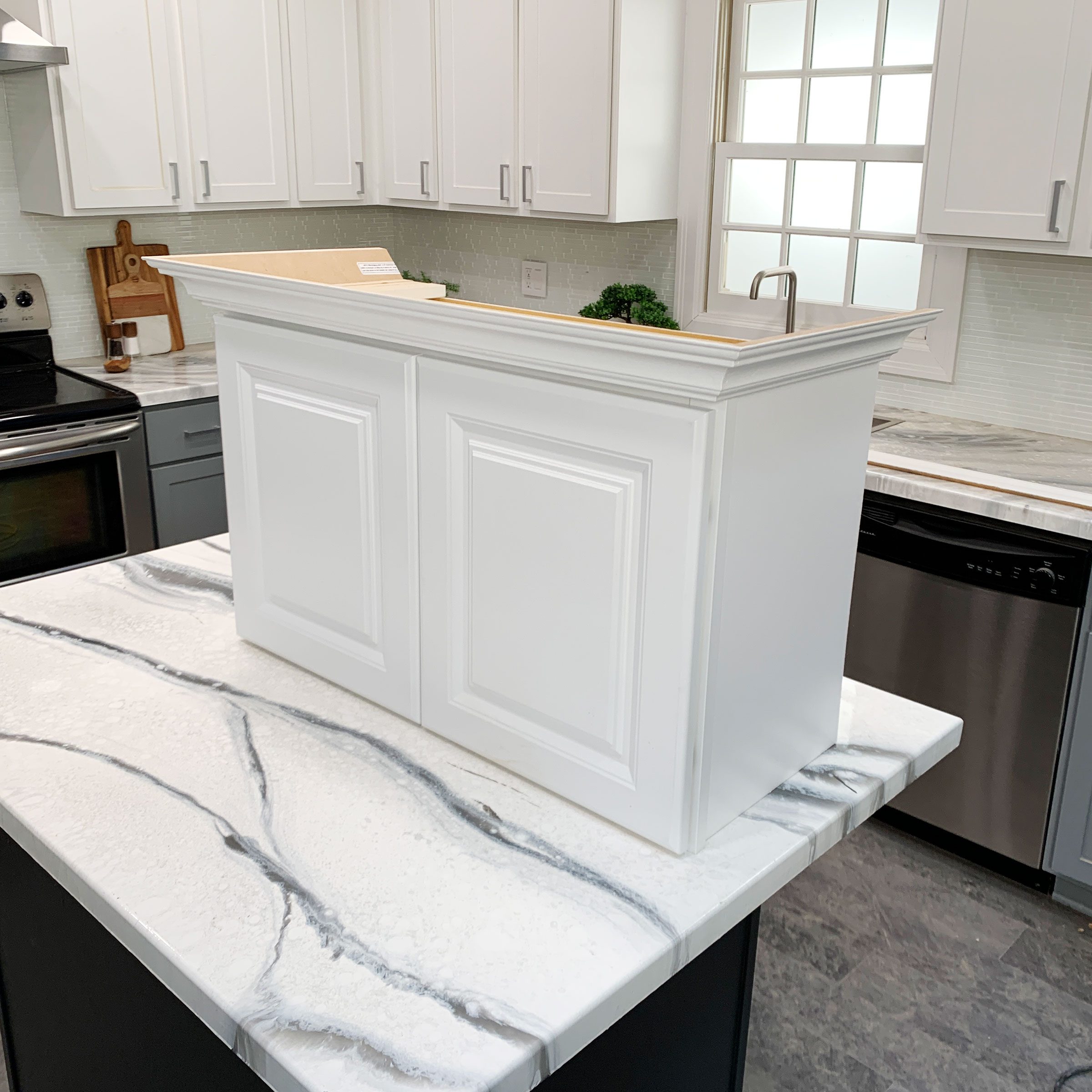 A white kitchen island stands on a marble countertop, featuring paneled sides and a top shelf, in a modern kitchen with cabinets and appliances.