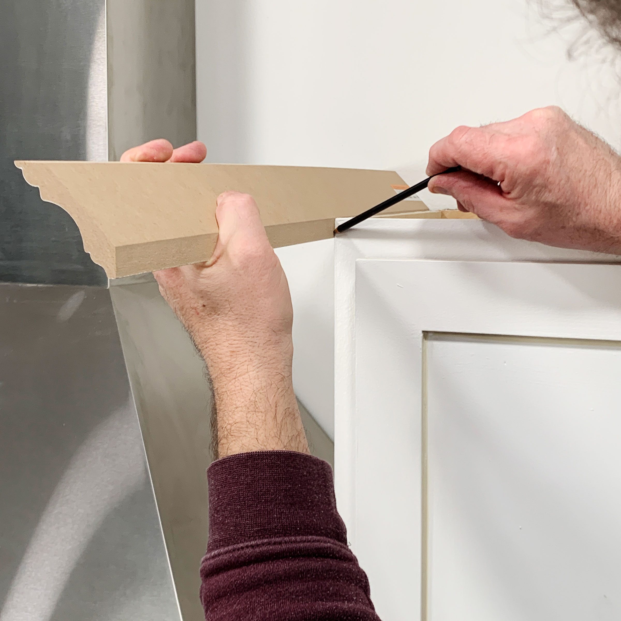 A person holds a decorative wood trim piece while marking its position against a white cabinet in a workshop setting.