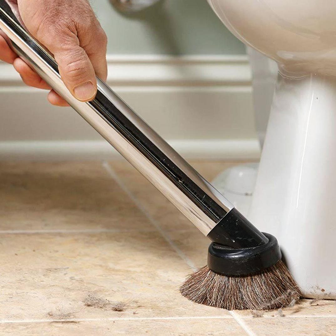 A hand holds a cleaning brush, removing dirt from the floor near a toilet in a bathroom setting. The surface is tiled with visible debris.