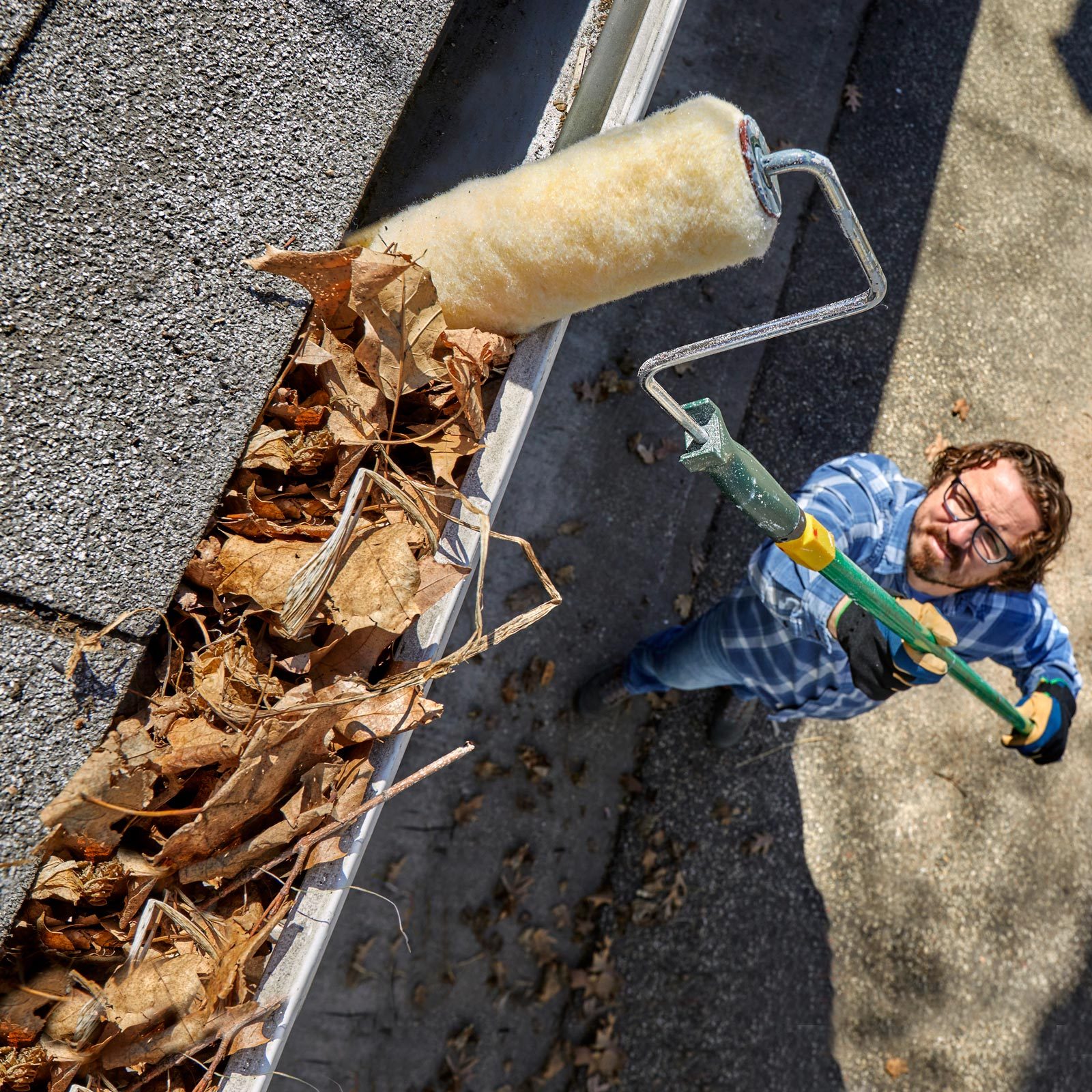 A man uses a tool resembling a paint roller to remove leaves from a rain gutter, with a residential setting in the background.