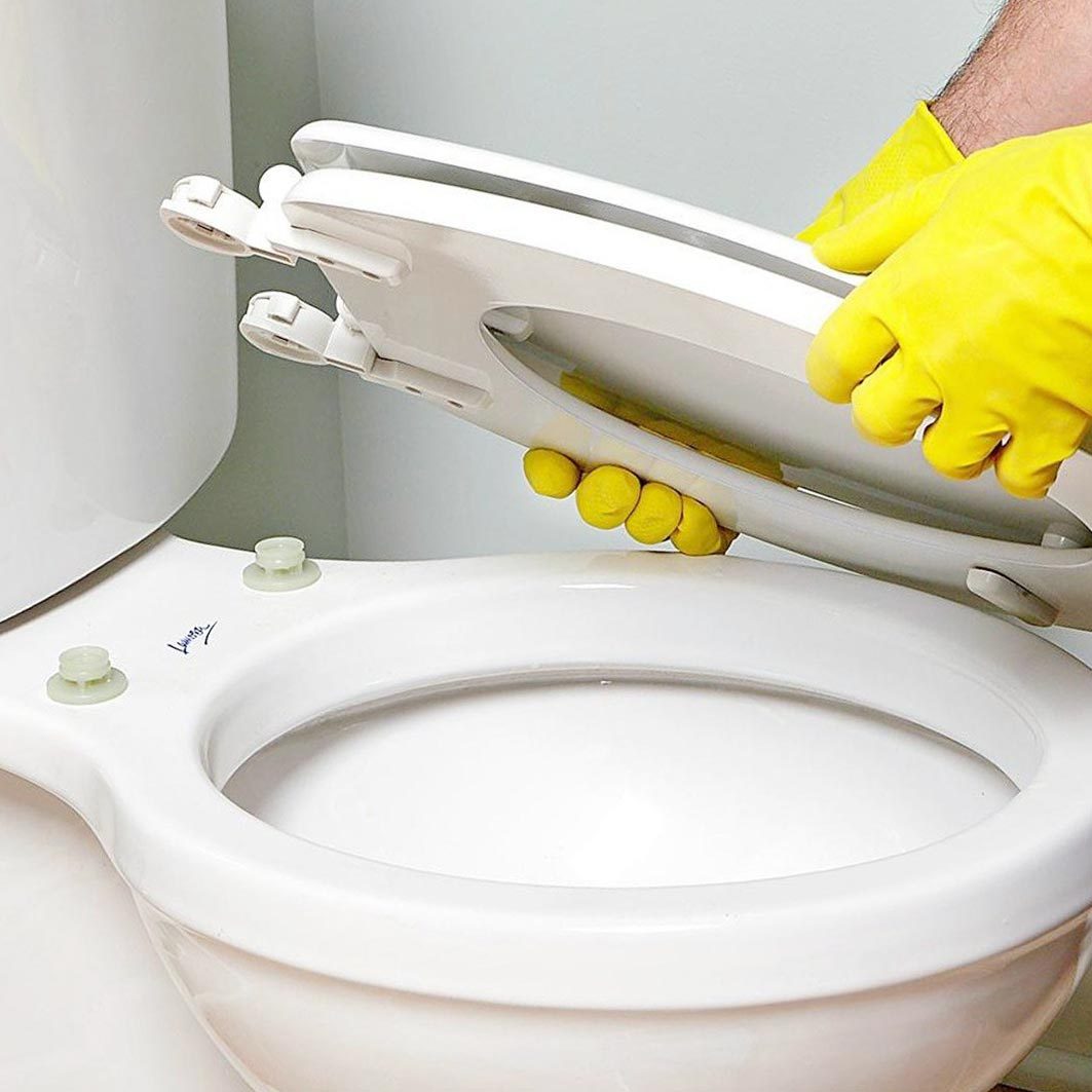 A person wearing yellow gloves lifts a toilet seat in a bathroom, revealing the white porcelain toilet bowl beneath it.