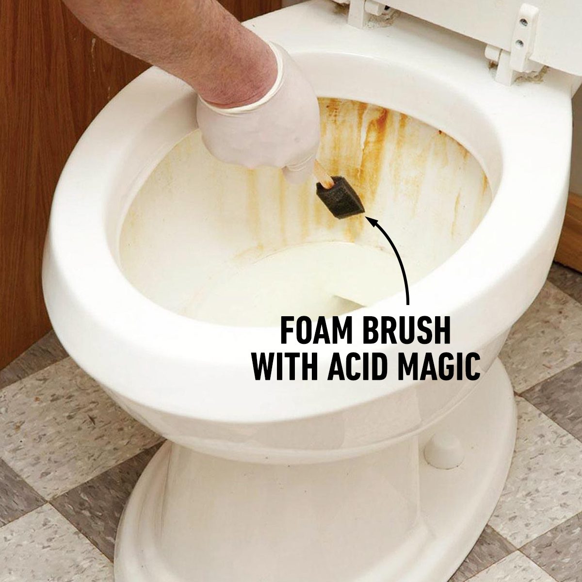 A hand wearing a glove scrubs a stained toilet bowl with a foam brush, while a clean floor and wooden wall are in the background.