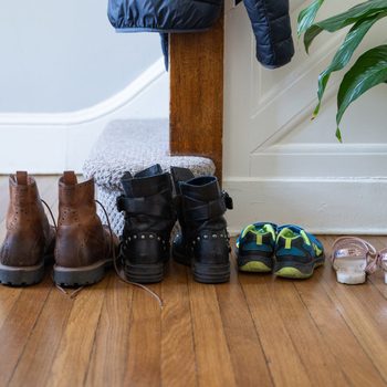 Four pairs of shoes are arranged neatly on a wooden floor by a staircase, with a jacket hanging nearby and a plant in the background.