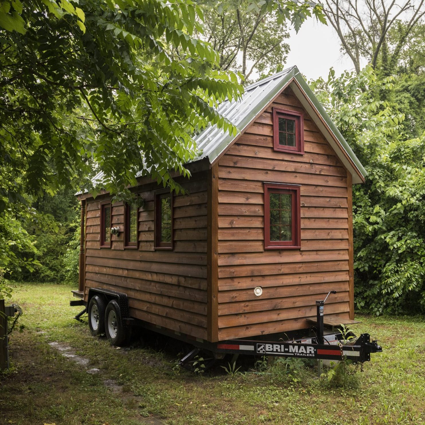 A wooden tiny house on wheels sits on overgrown grass, surrounded by lush greenery. Its peaked roof and red-framed windows create a rustic aesthetic.
