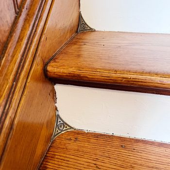 Wooden stair treads display decorative metal corner brackets, highlighting craftsmanship. The white wall provides a clean backdrop, accentuating the stairs' warm tones.