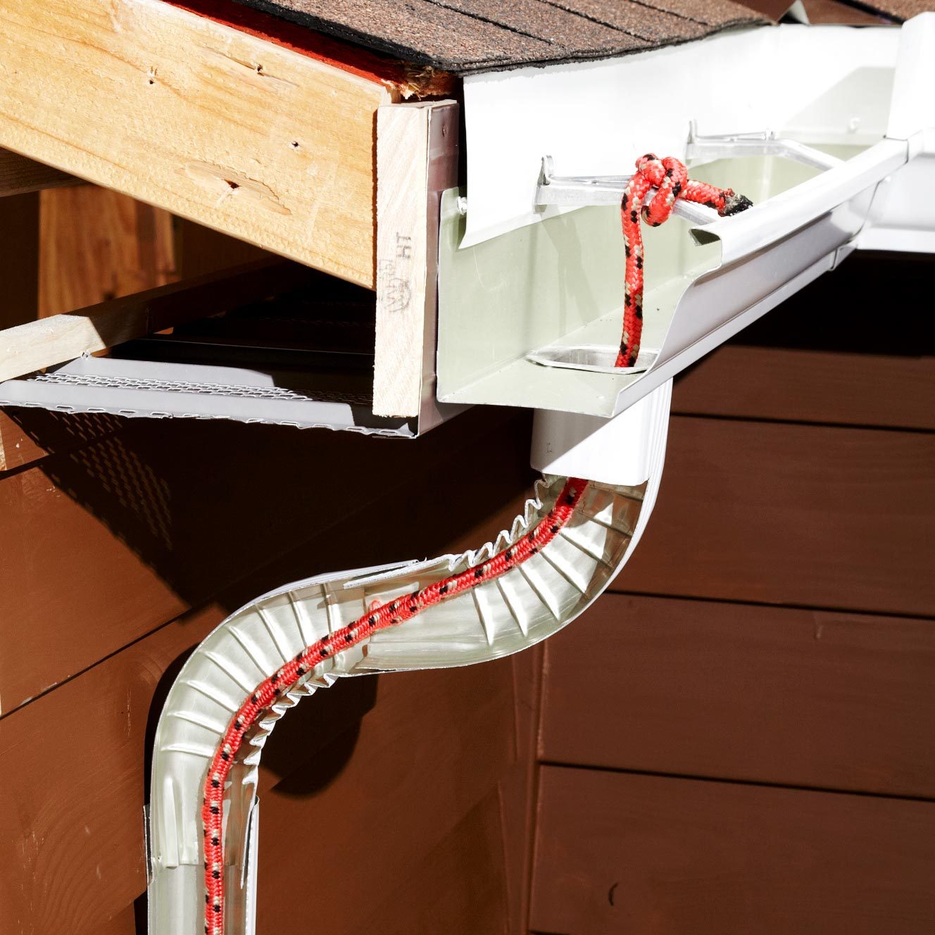 A gutter system channels water from a roof. A red rope is secured within the gutter, positioned against a wooden structure in sunlight.