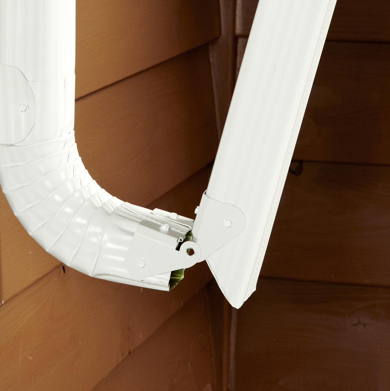 A white gutter system bends at a joint, directing water flow from the roof. It connects to a wooden wall, emphasizing a home maintenance function.